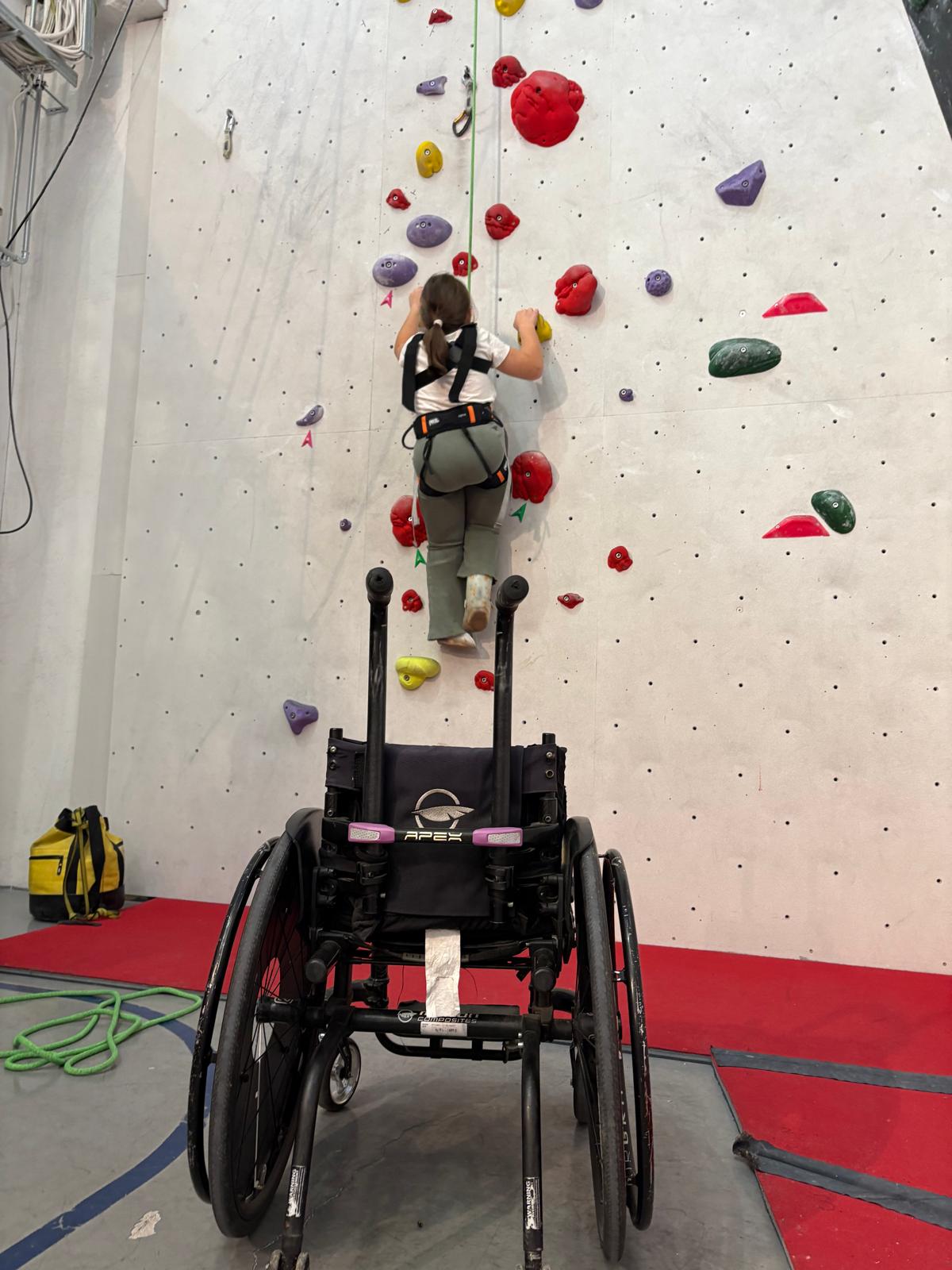 Kuvassa henkilö kiipeilee kiipeilyseinällä ja kuvan etualalla hänen pyörätuoli